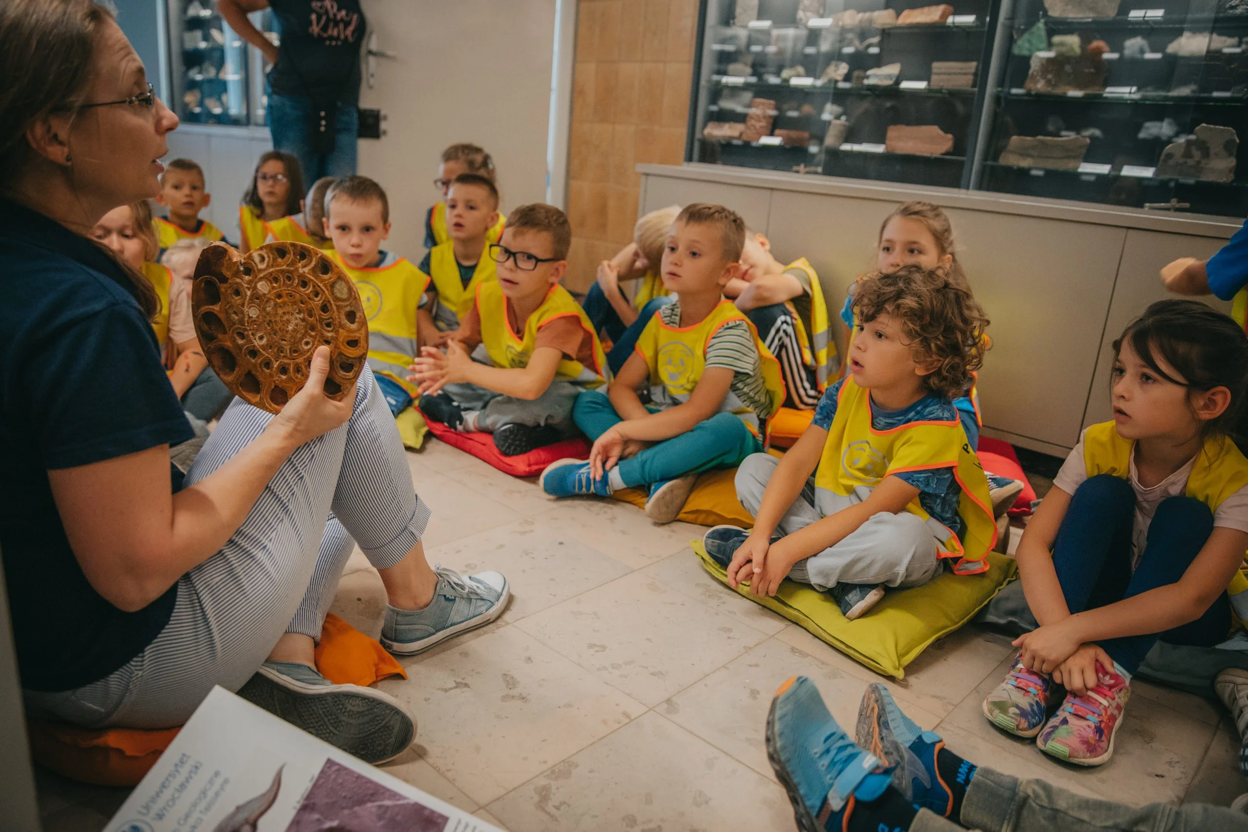 Na zdjęciu, edukatorka siedzi z lewej strony trzymając w ręce dużego amonita. Przed nią, na kolorowych poduszkach, siedzi zasłuchana grupa dzieci w wieku przedszkolnym.